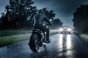 Motorradfahrer fährt bei Dunkelheut und Gewitter auf einer Landstraße, während Blitze den Himmel erleuchten und die Sicht stark eingeschränkt ist. Vor ihm ist ein Auto und eine gefährliche Situation.